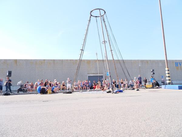 Eine dreibeinige Stahlpyramide / Trapezgerüst steht vor einem langgestreckten Gebäude und blauem Himmel. Rundherum Publikum.