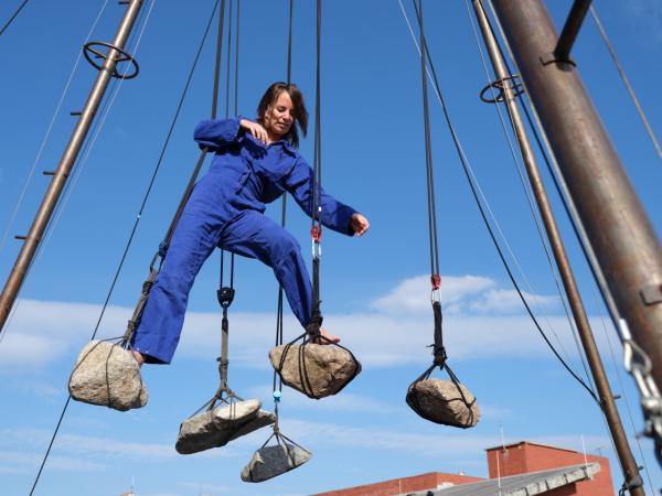 Jana Korb, eine weiße kurzhaarige cis Frau steht auf zwei hängenden Steinen. Im Blaumann vor blauem Himmel, in einem Stahlgerüst hängend.