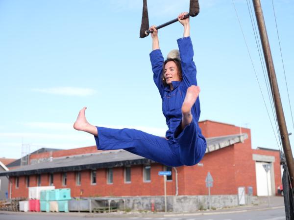 Jana Korb, eine weiße cis Frau, hängt an ihrem Trapez. Sie reckt ihre nackten Füße nach vorne und hat einen großen Stein auf dem Kopf. Sie ist mit einem blauen Blaumann bekleidet. Im Hintergrund ein rotes Ziegelgebäude vor hellblauem Himmel.