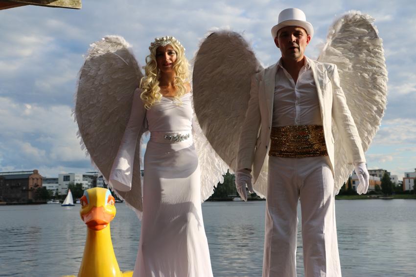 Zwei weiße Engel auf Stelzen stehen am Wasser, neben einer gelben Plastik-Ente.