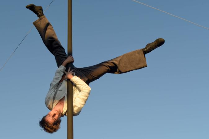 Anja Gessenhardt in der Rolle der Bühnenarbeiterin kopfüber an der Vertikalstange vor blauem Himmel.