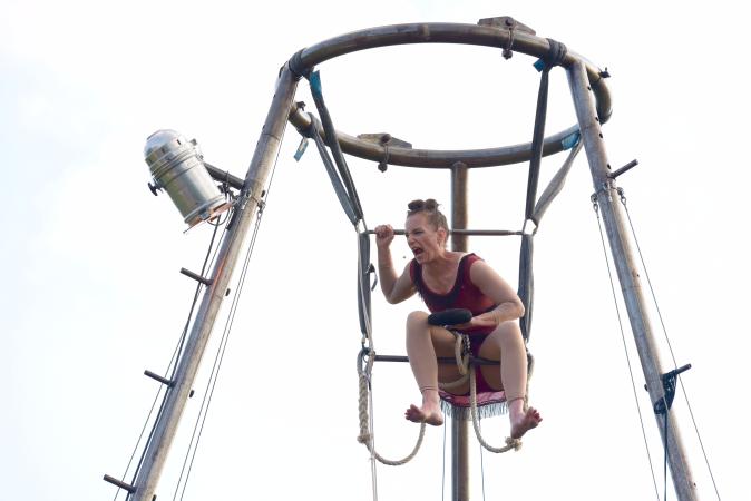 Jana Korb in der Rolle der Trapezkünstlerin ganz oben in ihrem Trapezgerüst vor weißem Himmel. Laut schreiend.