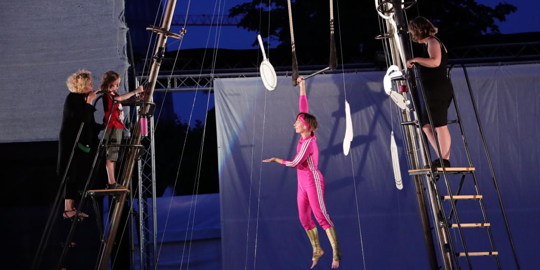 Jana Korb, eine weiße cis Frau, bekleidet mit einem pinken Overall, hängt einarmig an ihrem Trapez, das in ihrer dreibeinigen Stahlpyramide / Trapezgerüst hängt. Sie gestikuliert mit der freien Hand und unterhält sich mit drei Besucher*innen, die sie links und rechts auf Leitern in der Höhe besuchen. Es hängen ein paar stilisierte Alltagsobjekte aus Papier im Luftraum. Im Hintergrund sind Leinwände und Bäume zu sehen, sowie Himmel zur blauen Stunde.