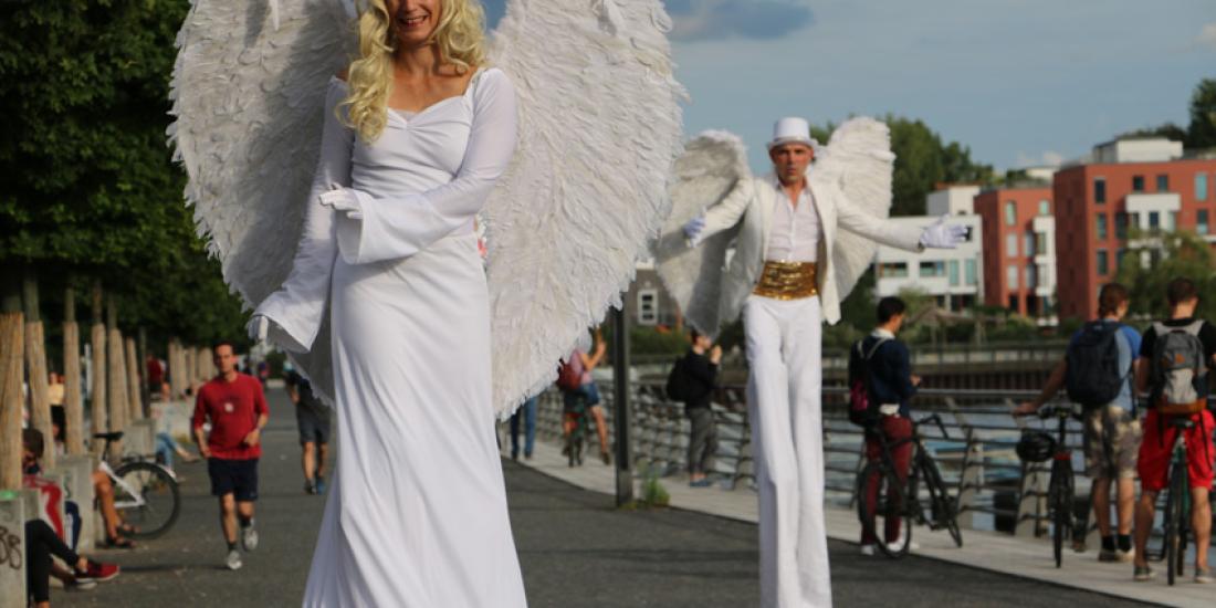 Zwei Stelzenläufer*innen als weiße Engel mit großen Flügeln auf einer Uferpromenade im Sommer.