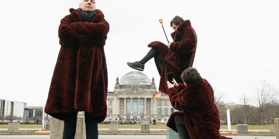 Drei Personen in weinroten Mänteln stehen vor dem Reichstagsgebäude. Einer trägt eine Krone, ein anderer tritt perspektivisch die Reichstagskuppel ein, während die dritte Person ein Foto davon macht.