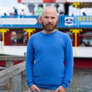 Junger Mann mit blauem Pullover am Hafen.