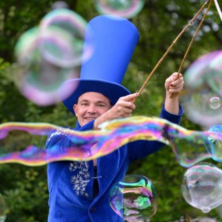 Ein blau gekleideter cis Mann mit Riesenhut macht hunderte von Seifenblasen.