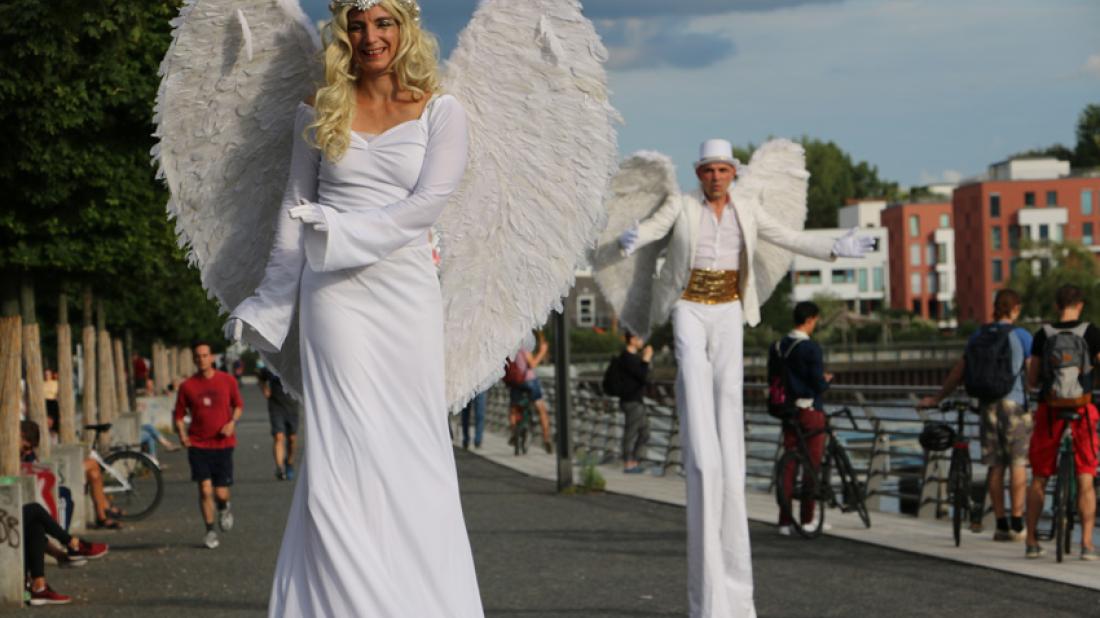 Zwei Stelzenläufer*innen als weiße Engel mit großen Flügeln auf einer Uferpromenade im Sommer.