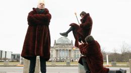 Drei Personen in weinroten Mänteln stehen vor dem Reichstagsgebäude. Einer trägt eine Krone, ein anderer tritt perspektivisch die Reichstagskuppel ein, während die dritte Person ein Foto davon macht.